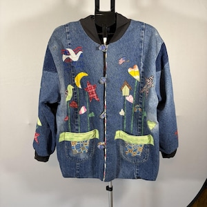 May include: A blue denim jacket featuring a black collar and cuffs, fastened with four patterned buttons. The front is decorated with colorful fabric appliqués including a white dove, a yellow crescent moon, a red star, a birdhouse, and hearts.
