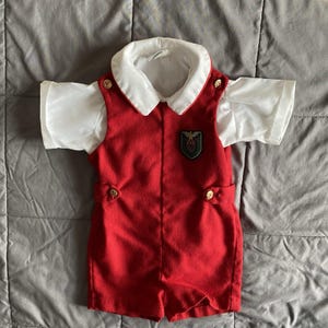 May include: A red romper with a white collared shirt and short sleeves. The romper has gold buttons and a green and gold emblem on the chest. The outfit is designed for a child.