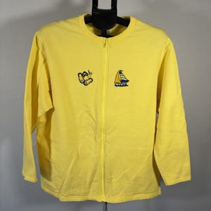 May include: A bright yellow zip-up sweatshirt with long sleeves. The sweatshirt features embroidered designs of a butterfly and a sailboat. The zipper runs down the center of the garment. The background is a neutral gray.