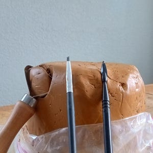 Pode incluir: Um bloco de argila castanha com ferramentas de escultura, incluindo uma ferramenta de laço com cabo de madeira e duas ferramentas com cabo preto. Uma pequena figura é visível no canto inferior esquerdo. A argila está num saco plástico transparente, sugerindo um projeto de artesanato ou arte.