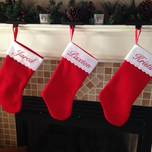 May include: Three red Christmas stockings with white trim and embroidered names, "Jared", "Paxton", and "Kristin", hanging on a white mantel above a fireplace.