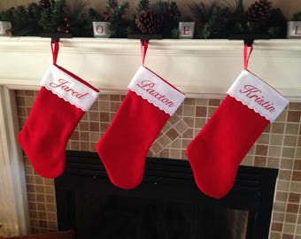 Puede incluir: Tres calcetines de Navidad rojos con ribete blanco están colgados en una repisa de chimenea blanca sobre una chimenea. Los calcetines están bordados con los nombres "Jared", "Paxton" y "Kristin".