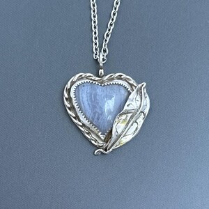 May include: Silver-toned heart-shaped pendant necklace. The pendant features a light blue stone set within a braided silver frame. A silver leaf detail adorns the side. The necklace hangs on a silver chain.