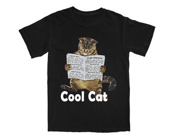 Pode incluir: Camiseta preta com um gato malhado lendo um jornal intitulado "Cat News". O gato está sentado e o texto "Cool Cat" está impresso em branco abaixo. É uma peça de roupa casual.
