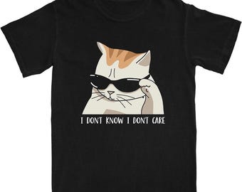 Könnte beinhalten: Schwarzes T-Shirt mit einer Katze mit Sonnenbrille und dem Text "I DON'T KNOW I DON'T CARE". Die Katze hat ein weißes Gesicht mit orangefarbenen und braunen Markierungen. Das T-Shirt ist aus weichem Material.