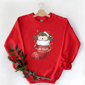 May include: A red sweatshirt featuring a Christmas-themed design. The graphic shows a cute hedgehog wearing a Santa hat and nestled in a Christmas stocking. The stocking is red with white snowflake patterns and trim.