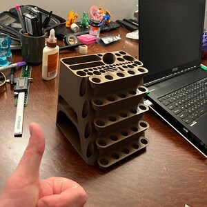 May include: A brown, multi-tiered organizer with various sized holes and compartments. The organizer is designed to hold art supplies, tools, or other small items. A laptop and other items are in the background.