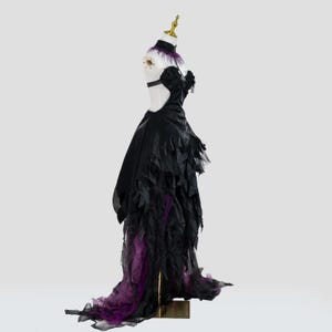 May include: A black formal gown with a dramatic design, featuring a high collar with purple feathers, a cut-out back, and a layered skirt with purple accents. The dress is displayed on a mannequin.