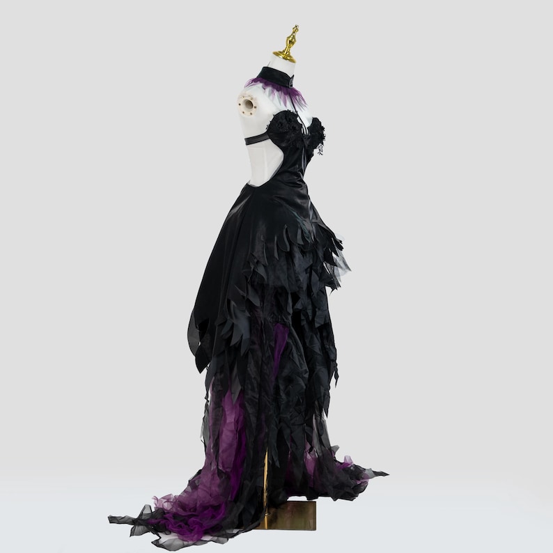 May include: A black formal gown with a dramatic design, featuring a high collar with purple feathers, a cut-out back, and a layered skirt with purple accents. The dress is displayed on a mannequin.