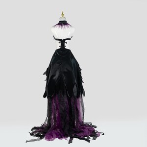 May include: A black and purple formal gown displayed on a mannequin. The dress features a black bodice with a high collar and a flowing skirt with black feather-like accents. The skirt transitions to a sheer purple fabric at the hem, creating a dramatic effect.