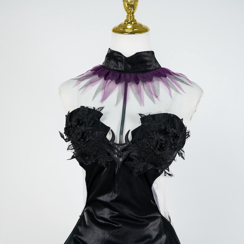 May include: A black satin dress with a high collar and a sheer, purple-trimmed neckline. The bodice features intricate black lace detailing. The dress is displayed on a mannequin with a gold-colored stand.