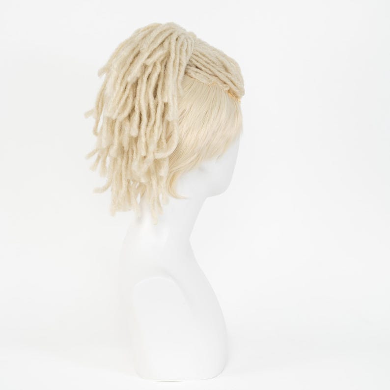 May include: A blonde wig with a dreadlock style, displayed on a white mannequin head. The wig features long, textured strands of light-colored hair, styled to one side. The mannequin is set against a plain white background.