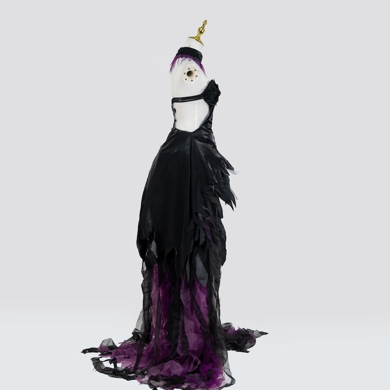 May include: A black and purple formal gown with a dramatic design. The dress features a fitted bodice with a cutout back and a flowing skirt with layered purple and black fabric. The gown is displayed on a mannequin.