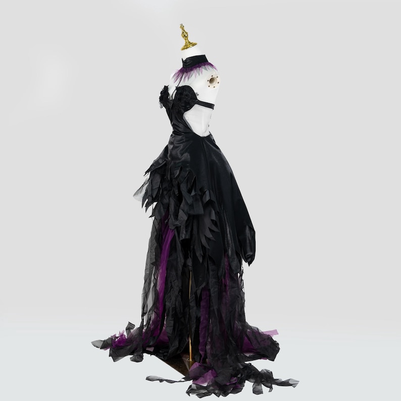 May include: A black formal gown with a high collar and open back, displayed on a mannequin. The dress features layered, feathery accents in black and purple, with a long train. The mannequin has a gold-colored headpiece.