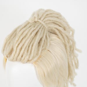 May include: A light blonde wig with a section of thick, textured dreadlocks tied at the crown. The dreadlocks are a lighter shade than the smooth, straight hair underneath. The wig is set against a plain white background.