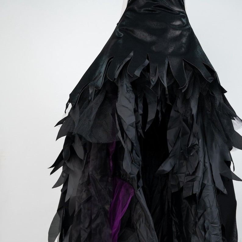 May include: A black costume dress with a layered design. The top portion has a jagged edge, and the skirt features multiple layers of fabric, including tulle and feather-like accents. A flash of purple fabric is visible.