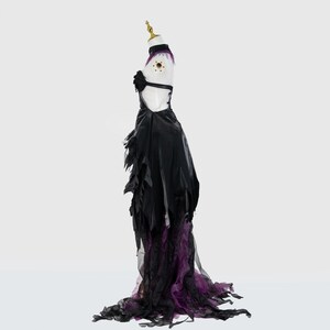 May include: A black and purple gothic-style dress on a mannequin. The dress features a halter top, a black skirt with tattered edges, and a flowing train with purple accents. The mannequin has a gold-colored head.