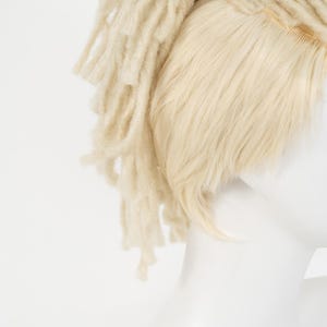 May include: A light blonde wig with dreadlocks and straight hair sections. The wig is on a white mannequin head. The dreadlocks are thick and textured, while the straight hair is smooth and fine. The wig is a light, neutral color.