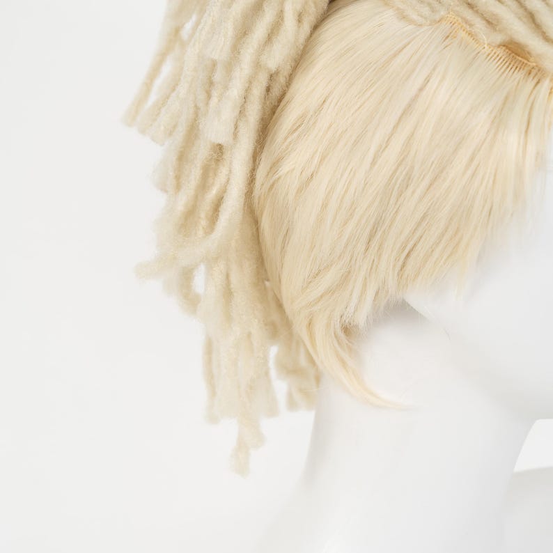 May include: A light blonde wig with dreadlocks and straight hair sections. The wig is on a white mannequin head. The dreadlocks are thick and textured, while the straight hair is smooth and fine. The wig is a light, neutral color.