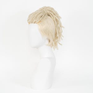 May include: A blonde wig with dreadlocks and a side part, displayed on a white mannequin head. The dreadlocks are a light beige color and are tied up at the crown of the head. The wig has a natural-looking hairline.