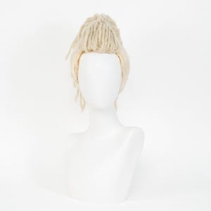 May include: A light blonde wig with dreadlock-style strands and a bun on top, displayed on a white mannequin head. The wig has a natural-looking texture and color, suitable for cosplay or costume use.