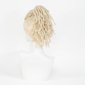 May include: A mannequin head displaying a light blonde wig with a section of cream-colored dreadlocks. The wig is styled with the dreadlocks gathered at the crown of the head. The mannequin is white, and the background is plain white.