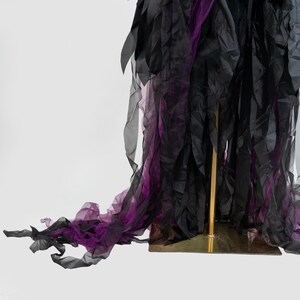 May include: A flowing, layered garment in shades of black and purple, displayed on a gold stand. The fabric appears to be sheer, with a textured, rippled effect. The design features long, trailing elements, creating a dramatic visual impact.