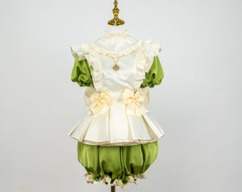 May include: A vintage-style outfit on a mannequin. The ensemble features a white satin top with ruffled edges and a cream-colored bow. The sleeves and bloomers are a vibrant green. A gold pendant hangs from the neckline.