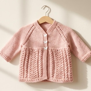 Puede incluir: Una chaqueta de punto de color rosa claro con mangas largas, escote en pico, mangas raglán y tres botones blancos en forma de mariposa en el centro. La parte inferior de la prenda presenta un patrón de punto calado texturizado.
