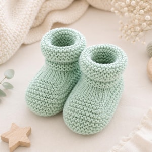 May include: A pair of light mint green baby booties featuring a chunky knit texture with a thick, folded cuff at the top. The booties have a rounded toe design and a consistent garter stitch pattern throughout the main body and the ankle collar.
