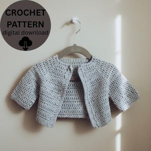 May include: A pale grey, crocheted baby cardigan hangs on a wooden hanger. The short-sleeved sweater is made of a textured stitch and has a single button closure. A circular graphic in the upper left corner reads "CROCHET PATTERN digital download".