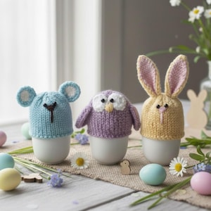 May include: Three knitted egg cozies in the shape of a mouse, owl, and bunny. The mouse cozy is light blue with white ears, the owl is purple with white and yellow details, and the bunny is yellow with pink ears. They sit on white egg cups.