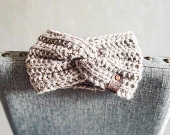 May include: A beige, chunky-knit headband with a twisted design. The headband is made of a thick, textured yarn and features a small, brown leather tag with a logo. The headband is resting on a gray chair.