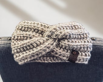 May include: A hand-knitted, gray and white twisted headband. The chunky knit design features a textured appearance. A small, dark brown leather tag is attached to the side. The headband is a cozy accessory.