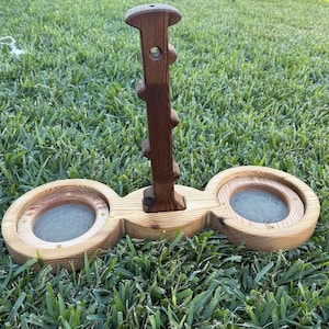 May include: A wooden bird feeder with a central vertical post and two circular feeding areas. The feeder is made of light-colored wood with a dark brown post and is set on a grassy lawn.