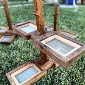 May include: Handmade wooden tiered plant stands with rectangular mesh-bottomed trays. The stands are made of natural-toned wood and are arranged on a bed of green grass. The design features a vertical support with horizontal platforms.