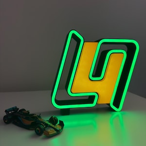 May include: A neon sign in the shape of the letter "L" and the number "4" with a yellow center and green outline. A small toy race car is in front of the sign. The sign is on a white surface.