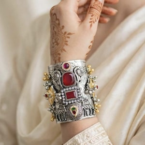 Può includere: Un ampio bracciale a fascia in argento con motivi animali in rilievo, lavorazioni in metallo ornate e diverse pietre preziose rosse incastonate. Il design include una pietra rossa rettangolare centrale, una pietra rossa rotonda sopra e piccoli accenti dorati.