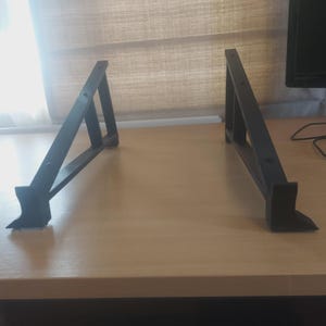 Pode incluir: Duas esquadrias de prateleira de metal preto são exibidas em uma superfície marrom clara. Cada esquadria tem um design triangular com um suporte vertical e uma base. As esquadrias são projetadas para suportar uma prateleira.