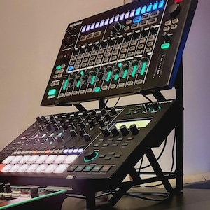 Peut inclure: Équipement de production de musique électronique noir avec plusieurs boutons, curseurs et boutons éclairés. L'équipement est monté sur un support en métal noir. L'unité supérieure porte le texte "Roland MX-1".