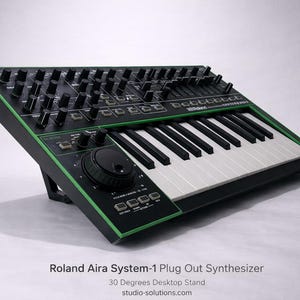 Pode incluir: Um sintetizador Roland Aira System-1 Plug Out preto e verde com teclado e inúmeros botões e controles deslizantes. O sintetizador está em um suporte de mesa de 30 graus. O teclado tem teclas pretas e brancas.