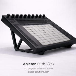 Op de afbeelding: Zwarte bureaustandaard voor Ableton Push 1/2/3, onder een hoek van 30 graden. De standaard heeft een raster van witte vierkanten en zwarte knoppen. De productnaam en website zijn onderaan zichtbaar.