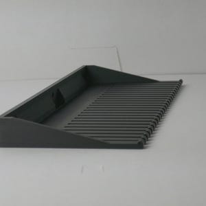 Puede incluir: Una bandeja gris oscuro en ángulo con una superficie de listones. La bandeja tiene forma rectangular con un borde elevado en un lado y una serie de líneas paralelas que forman la superficie. El artículo es de material sólido.