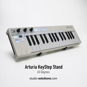 May include: A white Arturia KeyStep stand with black and white keys, knobs, and buttons. The stand is angled at 45 degrees and has the text "Arturia KeyStep Stand" and "studio-solutions.com" printed on it.