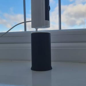 Op de afbeelding: Een witte Ring-beveiligingscamera gemonteerd op een zwarte cilindrische basis. De camera heeft een lens en het woord "Ring" erop gedrukt. De opstelling staat op een witte vensterbank, met een blauwe lucht zichtbaar door het raam.