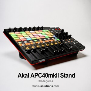 Puede incluir: Soporte negro Akai APC40mkII con un borde rojo. El soporte presenta una cuadrícula de botones, perillas y controles deslizantes multicolores. El texto "Akai APC40mkII Stand" se muestra debajo. El soporte está inclinado 30 grados.