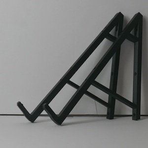 Puede incluir: Dos escuadras de estantería de metal negro. Cada escuadra tiene un diseño triangular con un brazo de soporte y un extremo en forma de gancho. Las escuadras están anguladas y parecen robustas, adecuadas para soportar estantes u otros objetos.