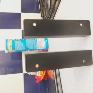 May include: Two black rectangular metal brackets with pre-drilled holes. A cylindrical container with a blue label is positioned between the brackets. The background features a checkered pattern of white and navy blue tiles.