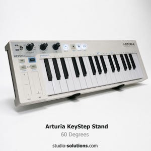 May include: A white Arturia KeyStep MIDI keyboard with black and white keys. The keyboard has several knobs and buttons, with the text "KeyStep" and "Arturia" visible. The stand is at a 60-degree angle.