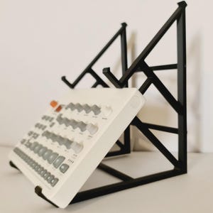 Puede incluir: Un dispositivo blanco de producción de música electrónica con perillas y botones grises se muestra en un soporte de metal negro. El soporte tiene un diseño geométrico y sostiene el dispositivo en ángulo, mostrando sus controles.